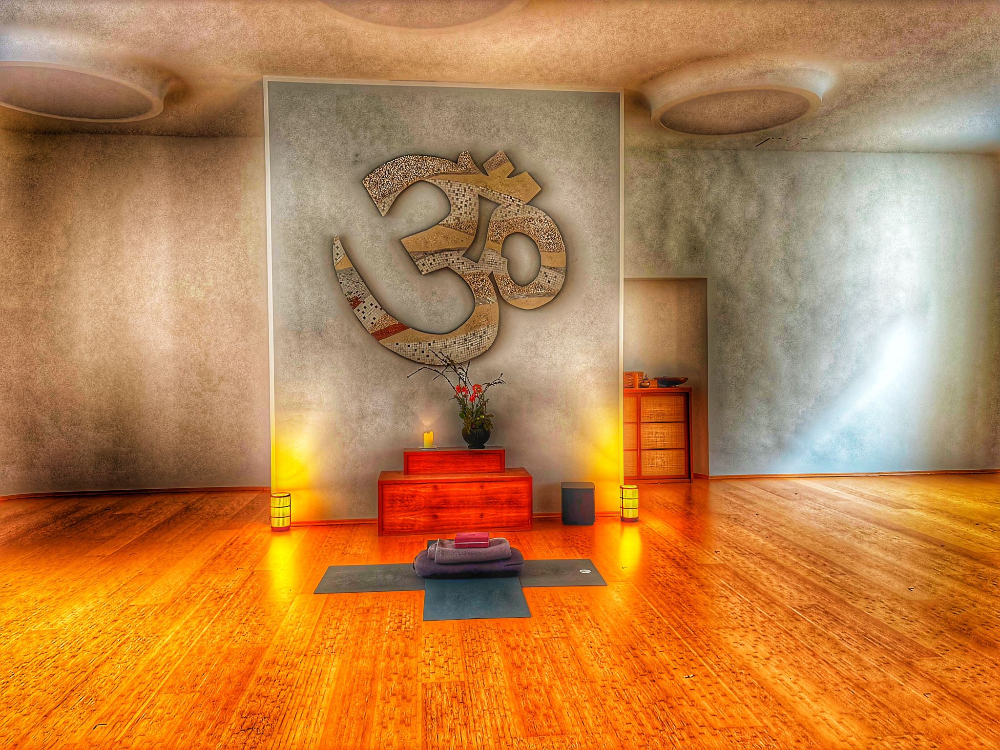 Großer Yogaraum im Spirit Yoga Charlottenburg, Bereich der Yogalehrerin mit Blume, kleinen Lampen, Lautsprechern und gekreuzten Yogamatten mit Zubehör